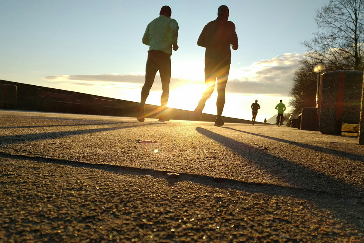 Two people running away from the camera with the sun shining in the background. Other runners can be seen in the distance on the same path.