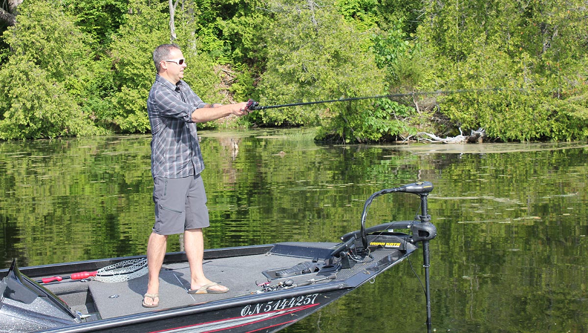 A man with a short-sleeved button-up shirt and shorts holds a fishing pole while standing on a boat in a river.