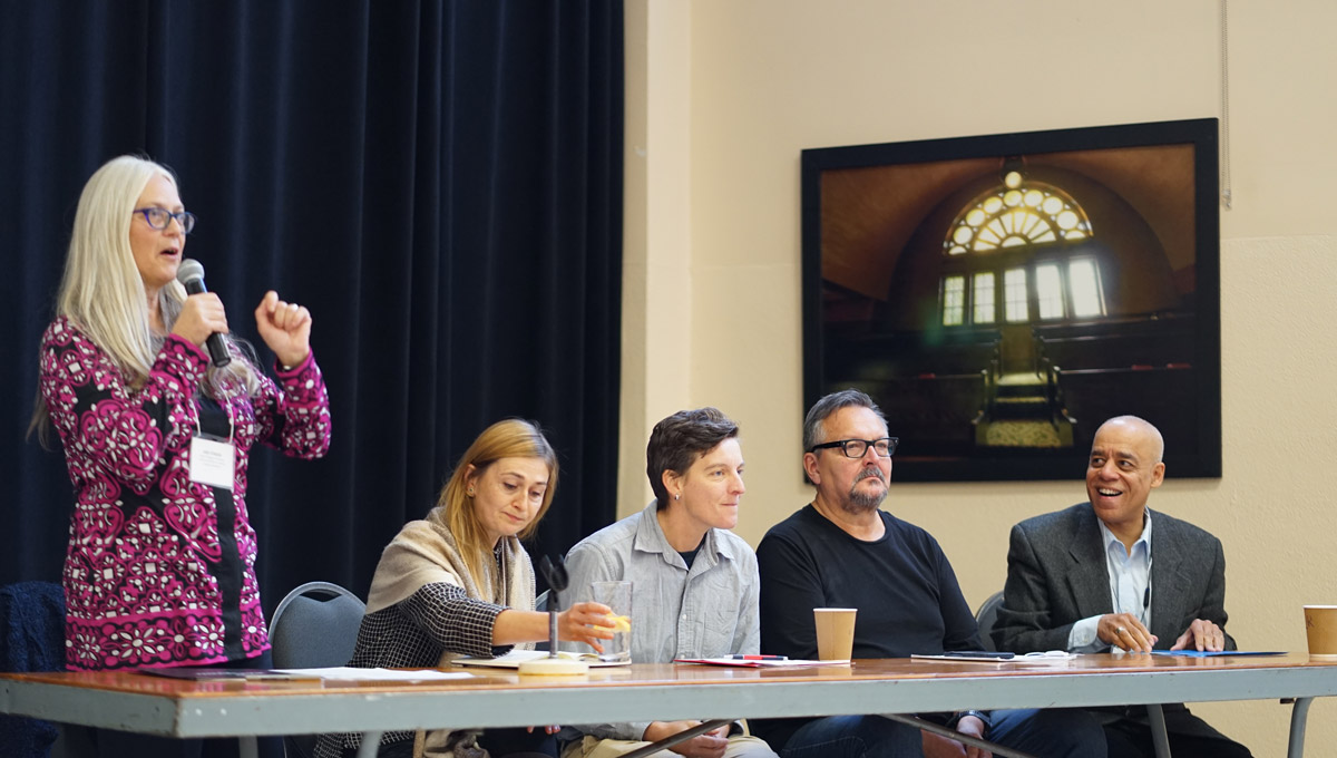 Jane Stinson speaks into a microphone while the members of a panel sit at a nearby table during the Work and Labour conference.