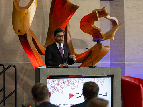 A man in a suit delivers a speech frrom a podium. An art exhibition can be seen in the background.